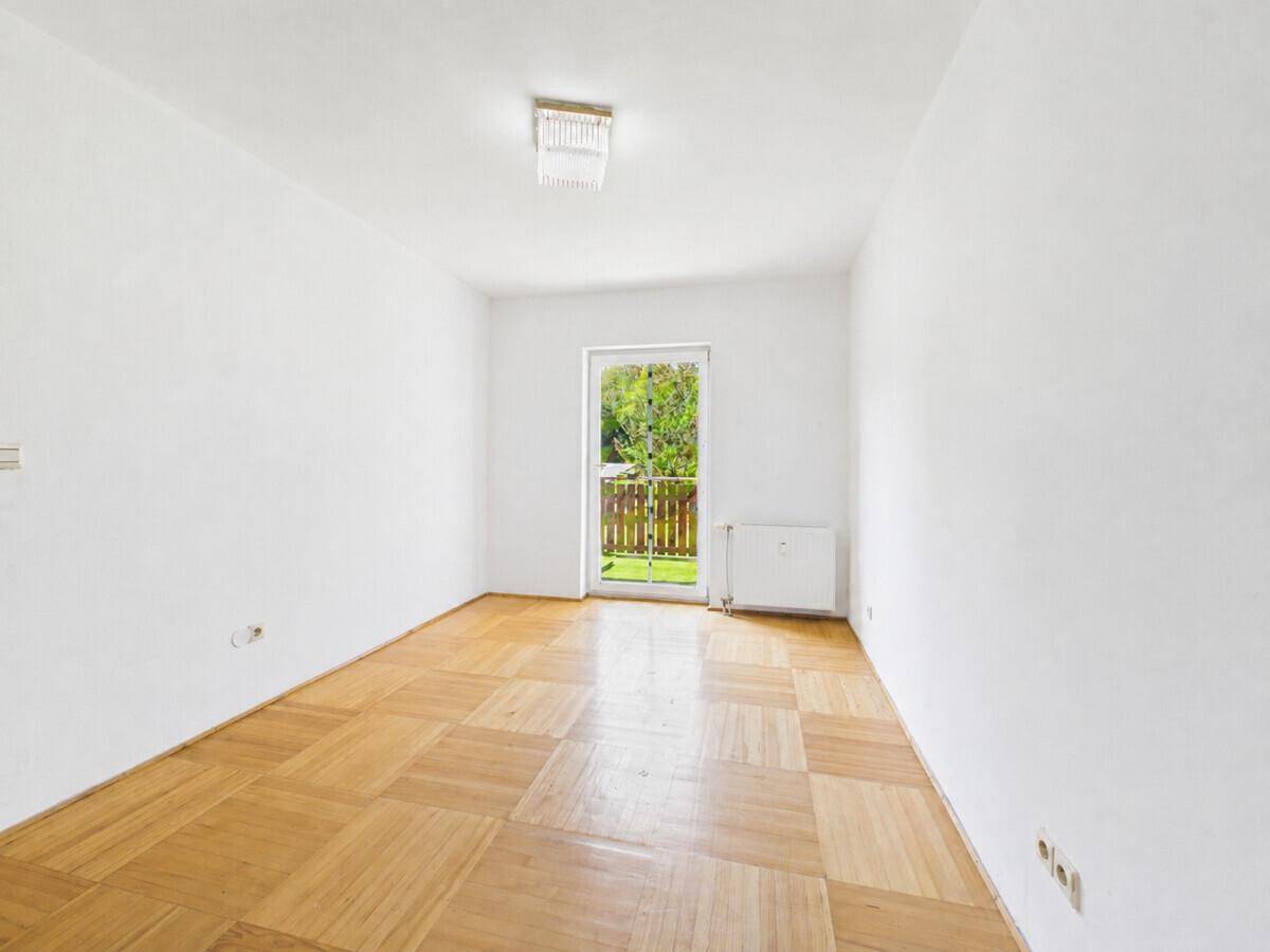 Helles Zimmer mit Parkettboden und Zugang zu einem kleinen Balkon mit Blick ins Grüne.