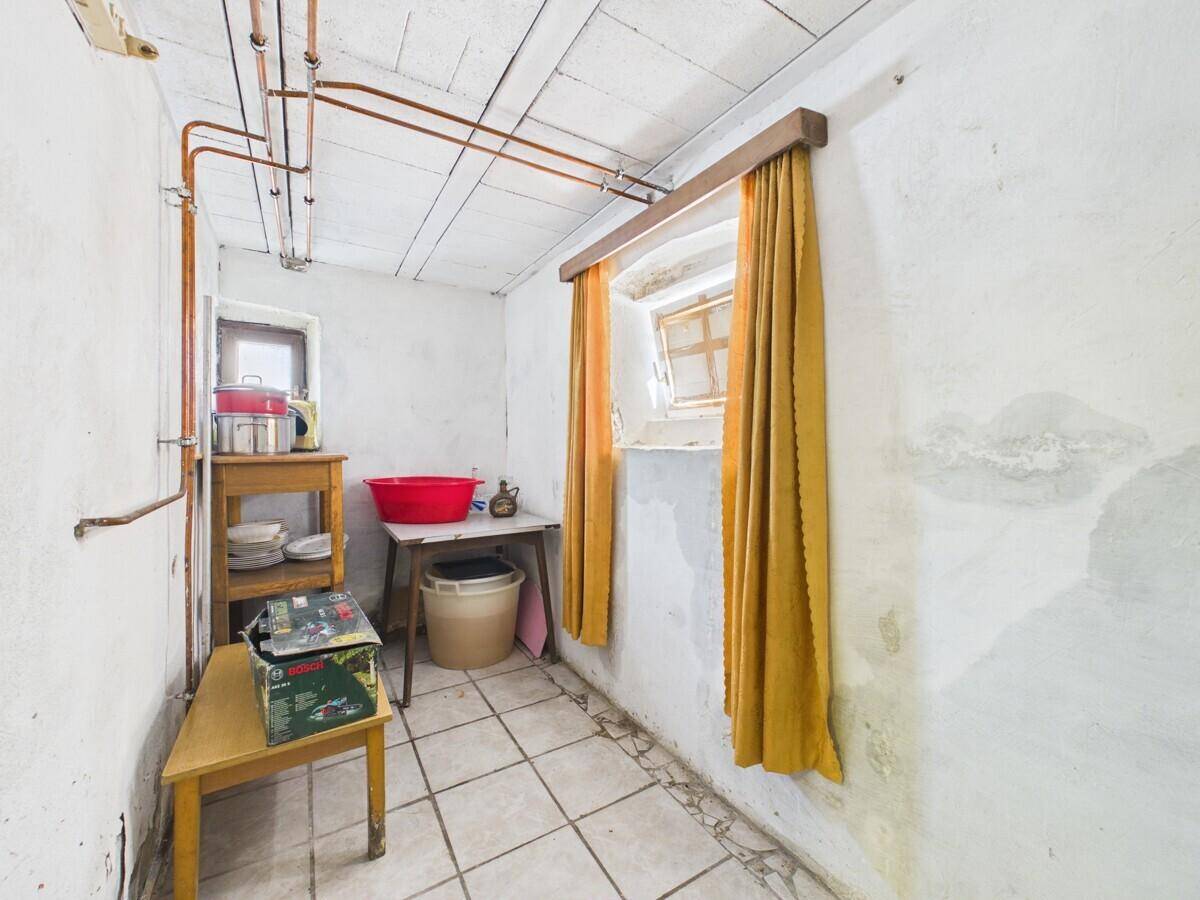 Einfacher Hauswirtschaftsraum mit Fliesenboden, sichtbaren Rohren und zwei kleinen Fenstern.