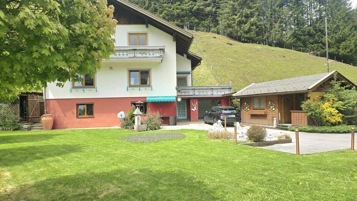 Haus mit Carport, Nebengebäude und großem Garten vor bewaldetem Hügel.