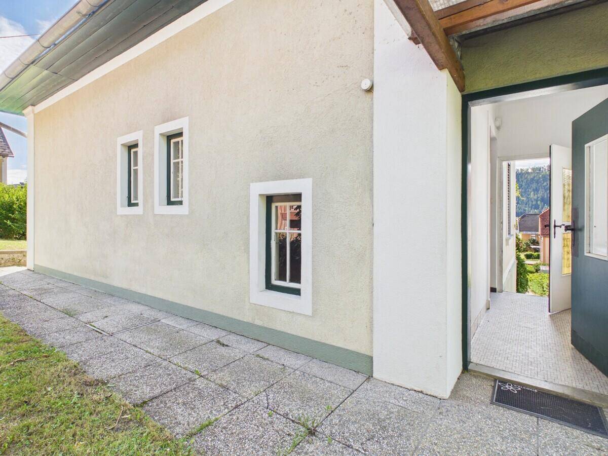 Hausfassade mit Eingangstür und kleinen Fenstern, umgeben von Grünflächen.