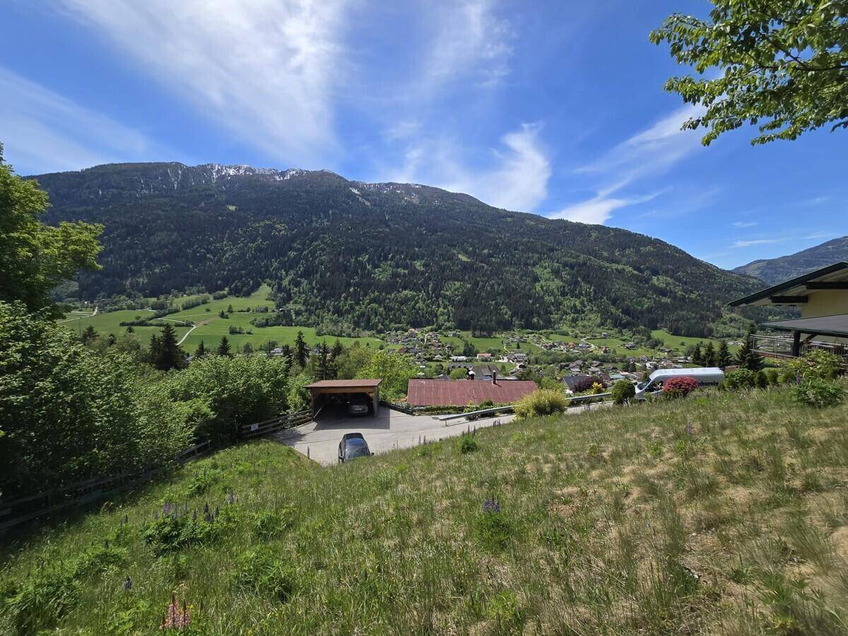 Weitläufiger Blick über eine grüne Wiese auf ein Dorf und die umliegenden Berge unter blauem Himmel.