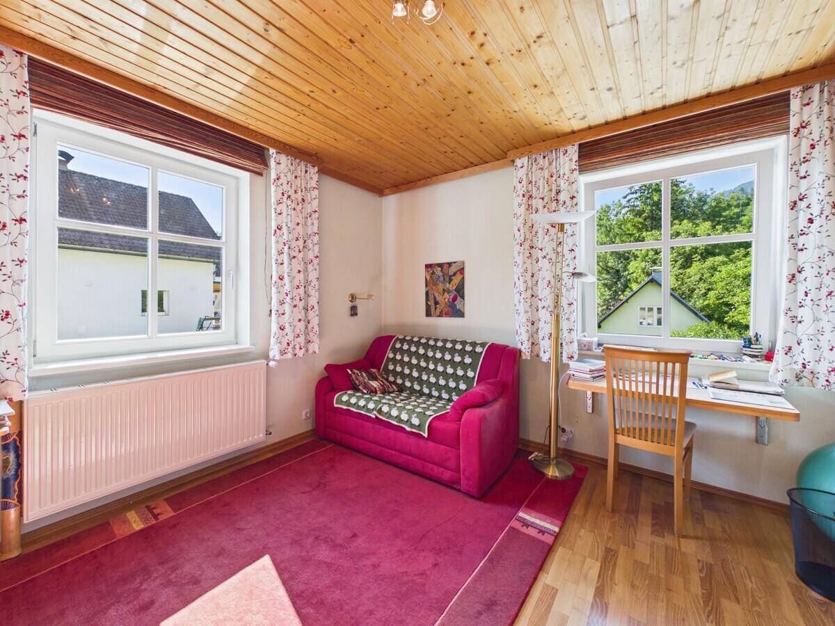 Helles Zimmer mit Holzbalkendecke, rotem Sofa und zwei Fenstern mit Blick ins Grüne.