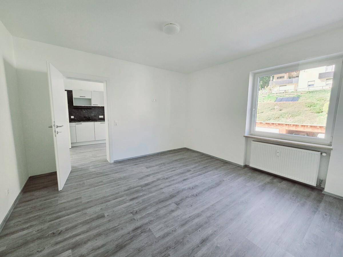 Geräumiges Zimmer mit dunklem Laminatboden, weiß gestrichenen Wänden und Heizkörper unter dem Fenster.