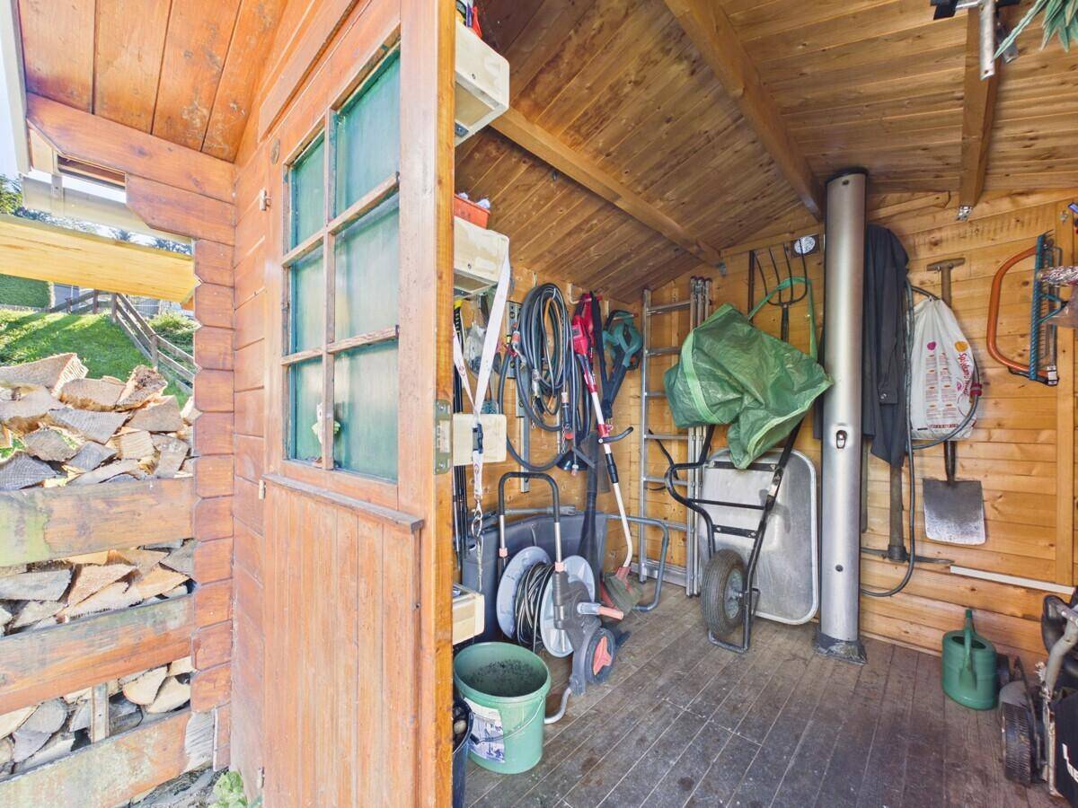 Innenansicht des Holzschuppens mit zahlreichen Gartengeräten, einer Schubkarre und gestapeltem Brennholz.