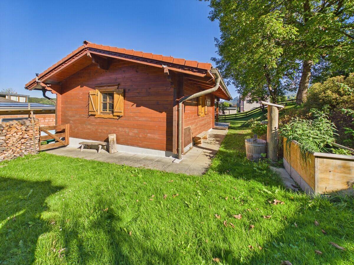 Seitenansicht des Holzchalets mit gestapeltem Brennholz, einer Holzbank und einem Hochbeet im Garten.