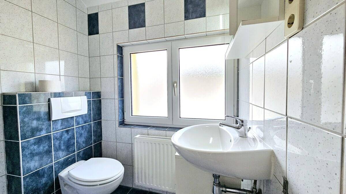 Helles Badezimmer mit Fenster, Toilette und Waschbecken, ausgestattet mit weißen und blauen Fliesen.