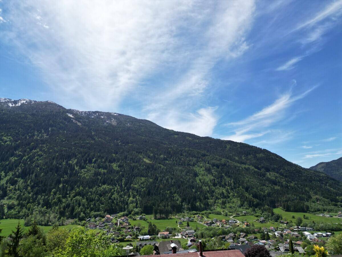 Breiter Blick auf eine bewaldete Bergkette und ein Dorf im Tal unter einem weiten blauen Himmel.