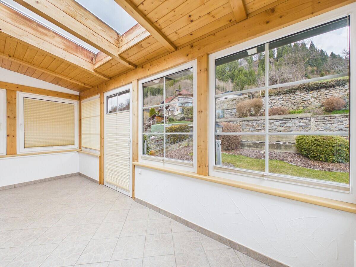 Heller Wintergarten mit Holzdecke, großen Fenstern und Blick auf die begrünte Hanglage.