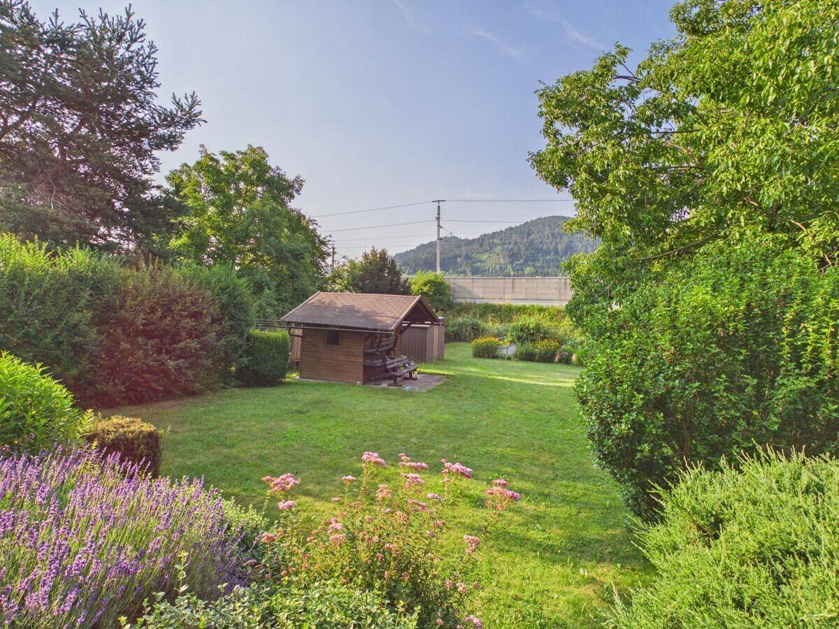 Gepflegter Garten mit Rasen, Blumenbeeten, Gartenhaus und Blick auf die bewaldeten Hügel.