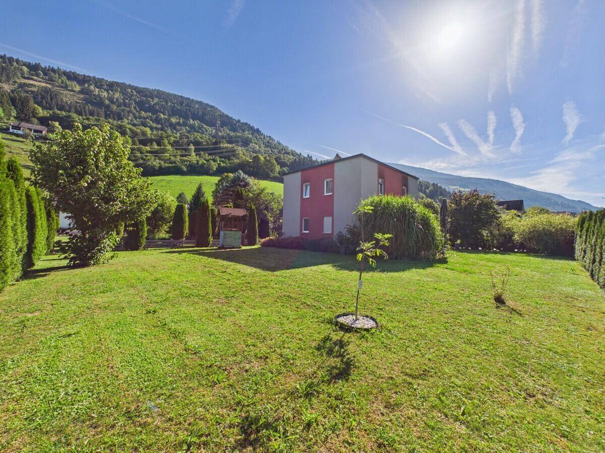 Sonnige Gartenansicht des Hauses mit gepflegtem Rasen und grüner Hügellandschaft im Hintergrund.