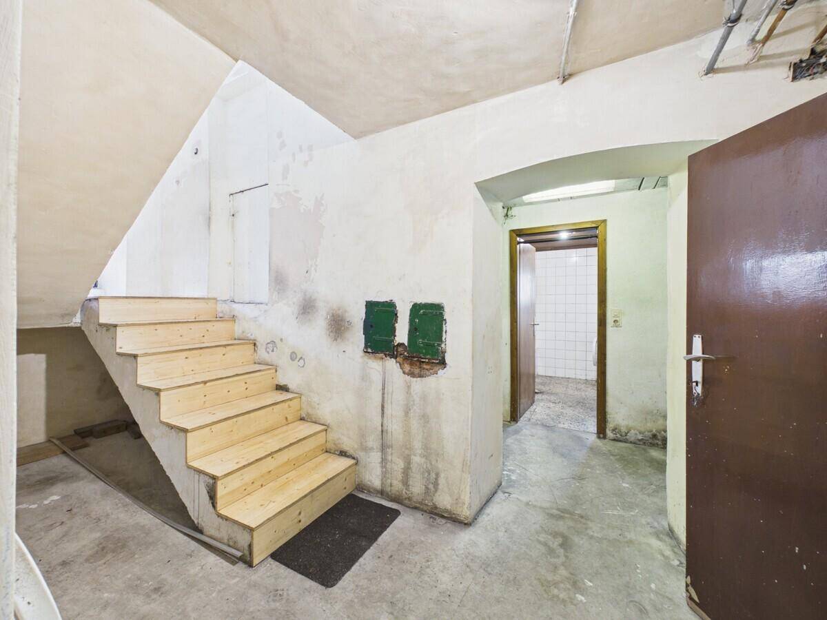 Kellerbereich mit Betonwänden, unfertiger Holztreppe und einem Durchgang zu einem weiteren Raum.