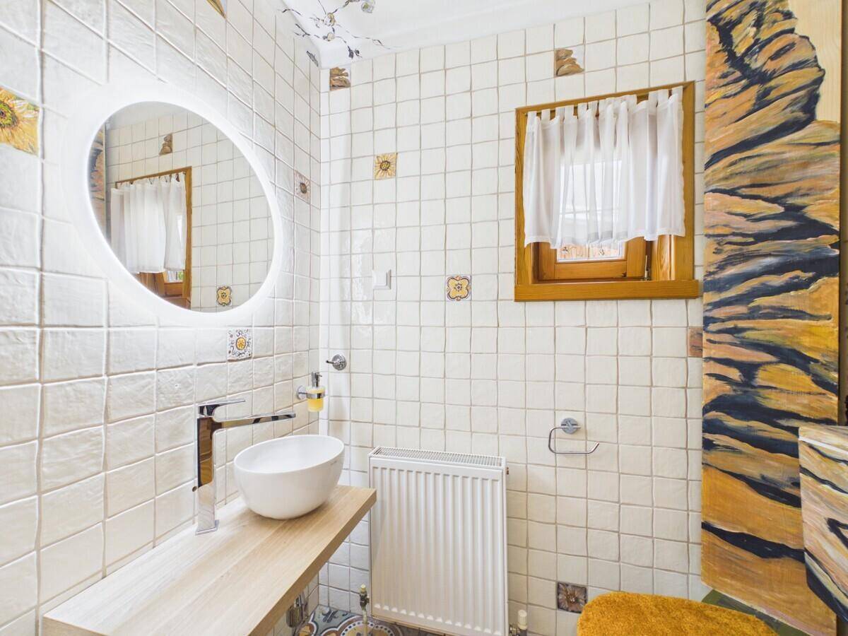 Helles Badezimmer mit kleinen weißen Fliesen, rundem beleuchtetem Spiegel und Fenster.