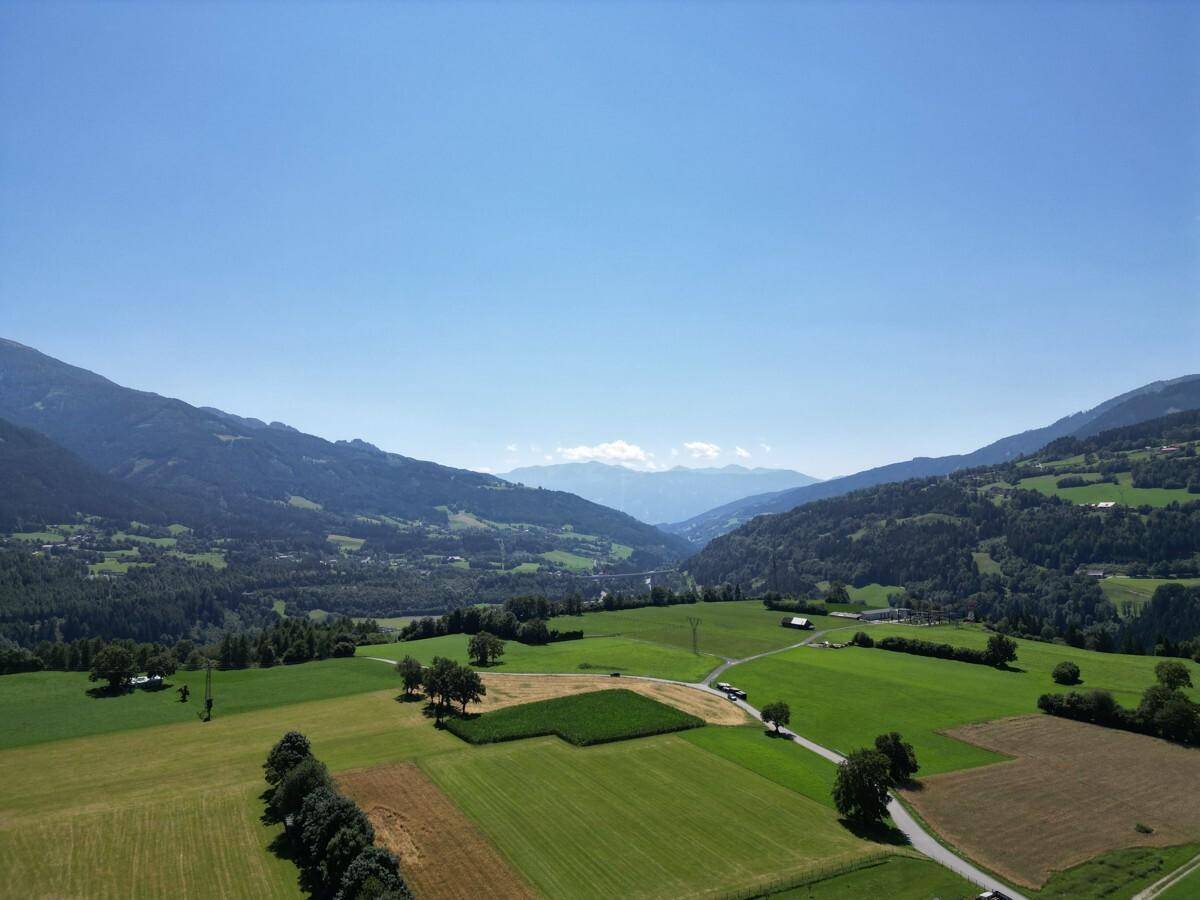 Weitläufige Luftaufnahme eines Tals mit grünen Feldern, Straßen und fernen Bergen unter einem strahlend blauen Himmel.