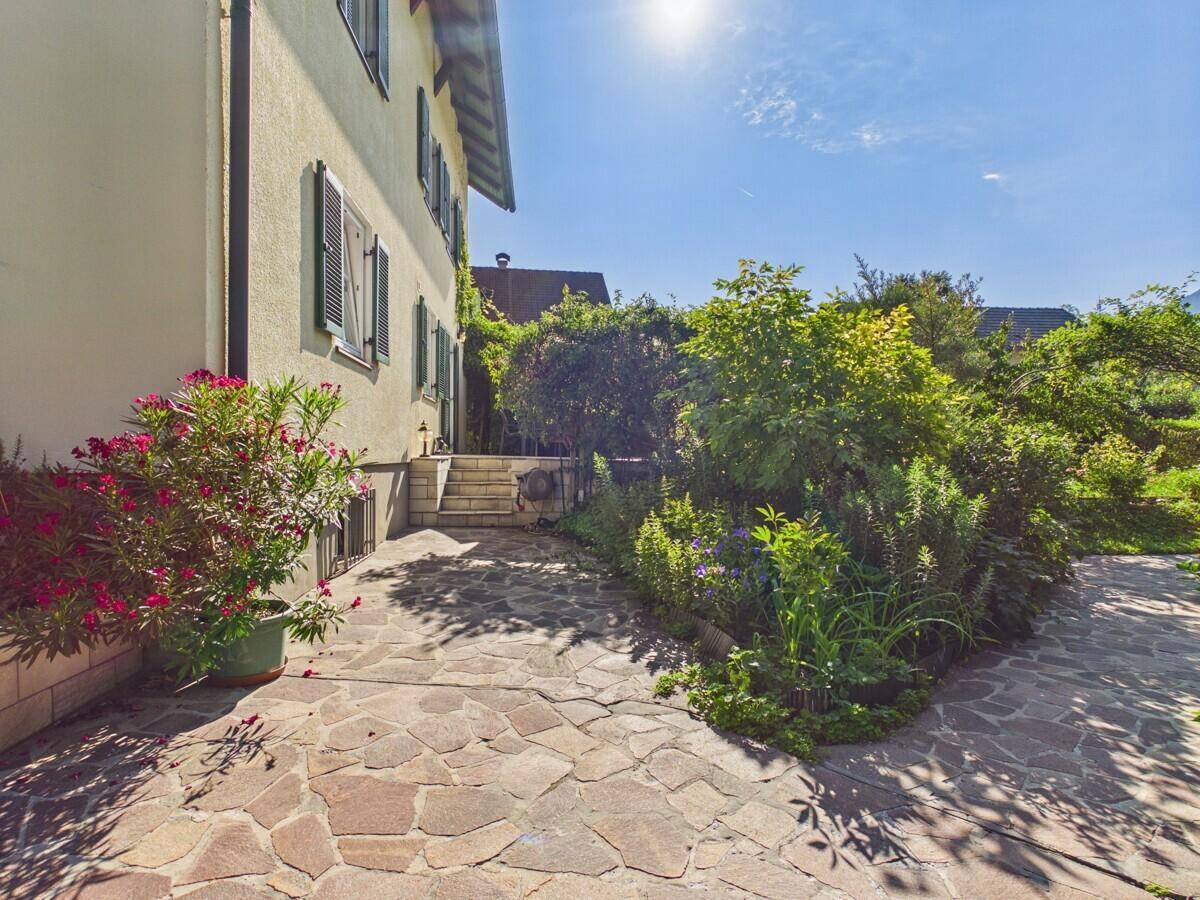Sonniger, gepflasterter Terrassenbereich neben dem Haus, umgeben von üppiger Bepflanzung.