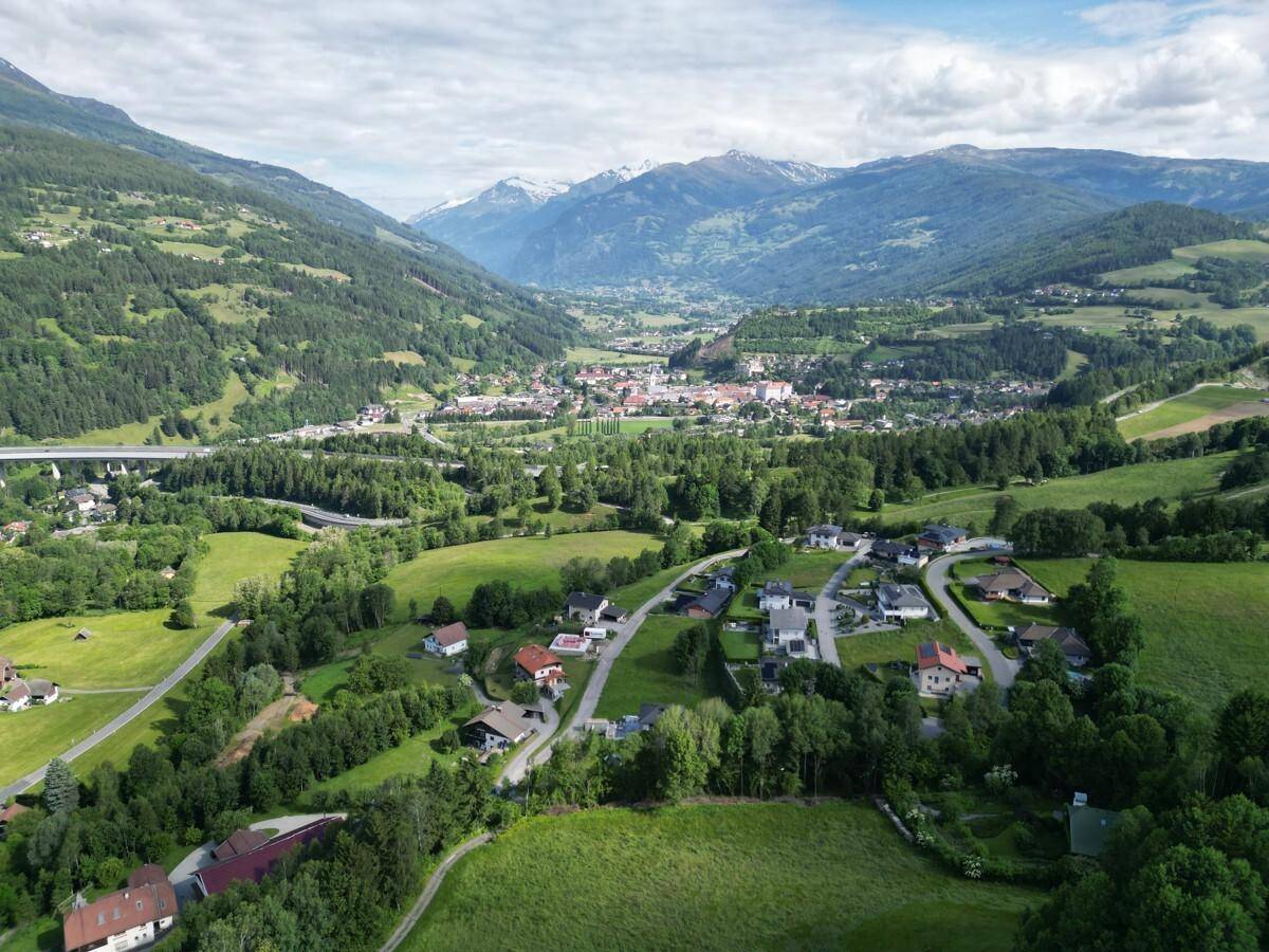 Beeindruckende Luftaufnahme eines Tals mit einer Stadt im Hintergrund, umgeben von grünen Bergen und schneebedeckten Gipfeln.