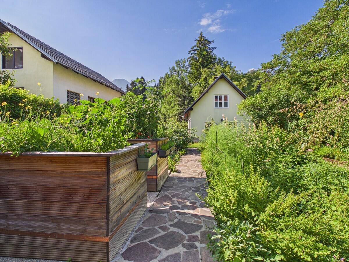 Gartenweg zwischen Hochbeeten mit Nutzpflanzen und Blick auf ein Nebengebäude.