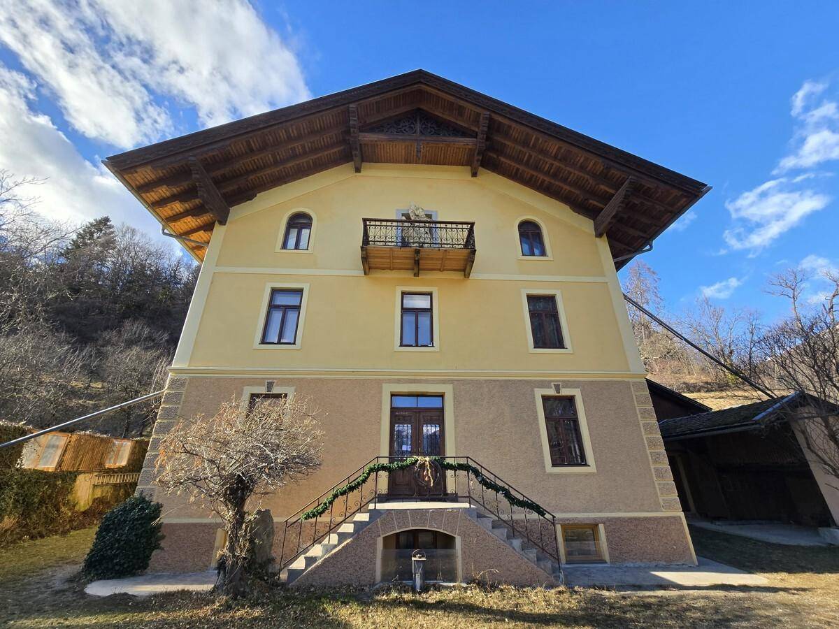 Traditionelles Gebäude mit gelber Fassade, Holzbalkonen und Satteldach in alpiner Umgebung.