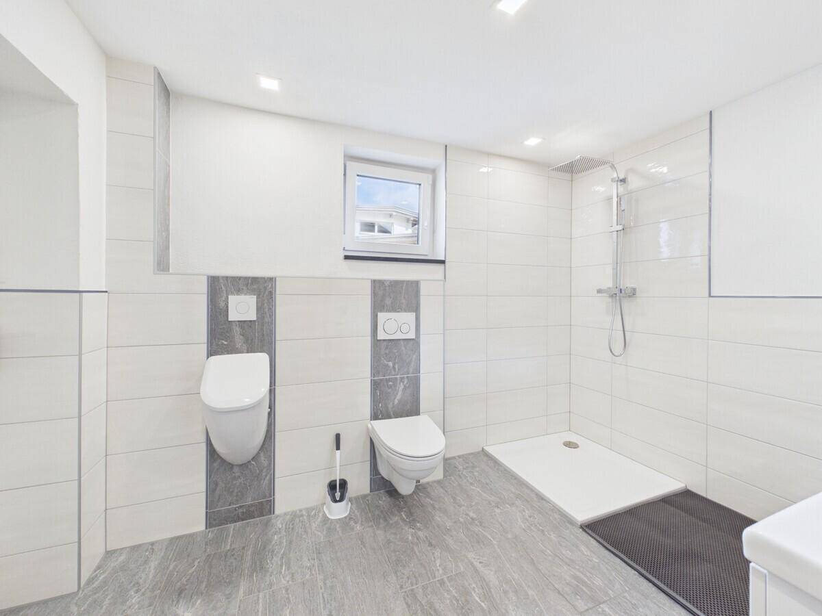 Helles Badezimmer mit ebenerdiger Dusche, WC, Urinal und Fenster.