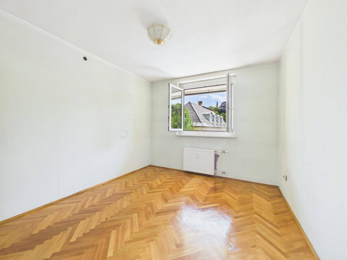 Leeres Zimmer mit Fischgrätparkett, weißen Wänden und Fenster mit Blick auf Dächer.