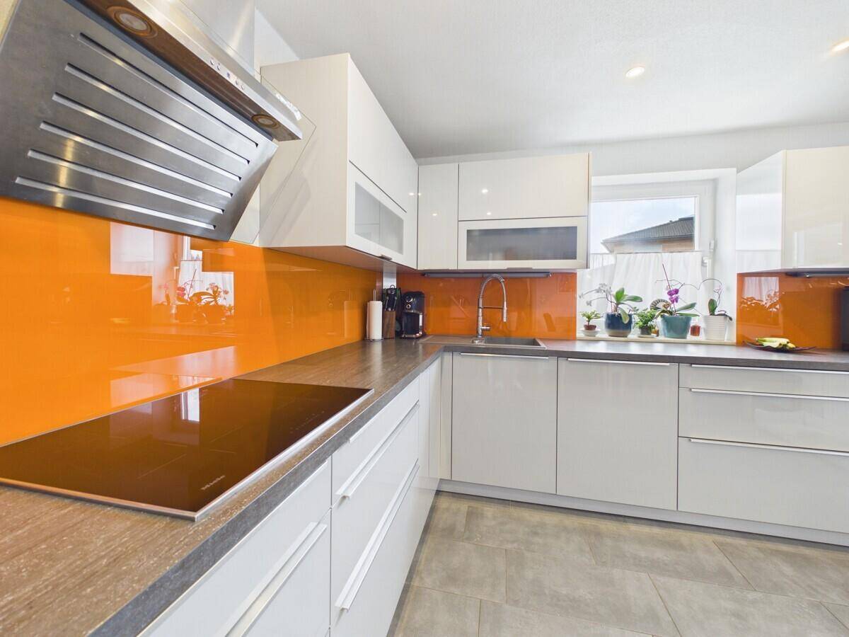 Helle Einbauküche mit Induktionskochfeld, orangefarbener Glasrückwand und Fenster.