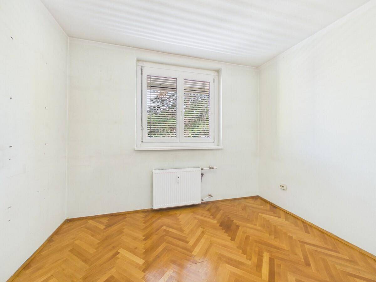 Helles, leeres Zimmer mit Fischgrätparkett, weißen Wänden und Fenster mit Jalousien.