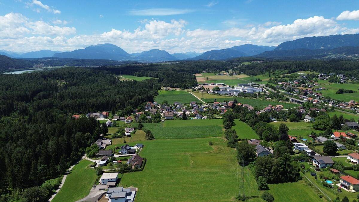 Weitläufige Luftaufnahme einer ländlichen Siedlung mit Feldern, Wäldern und einer Bergkette im Hintergrund.