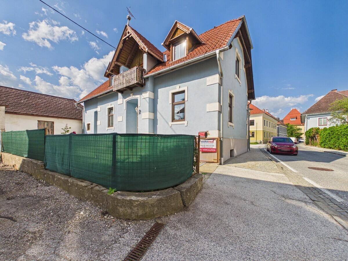 Außenansicht des hellblauen Hauses von der Straße aus mit einem "Zu verkaufen"-Schild.
