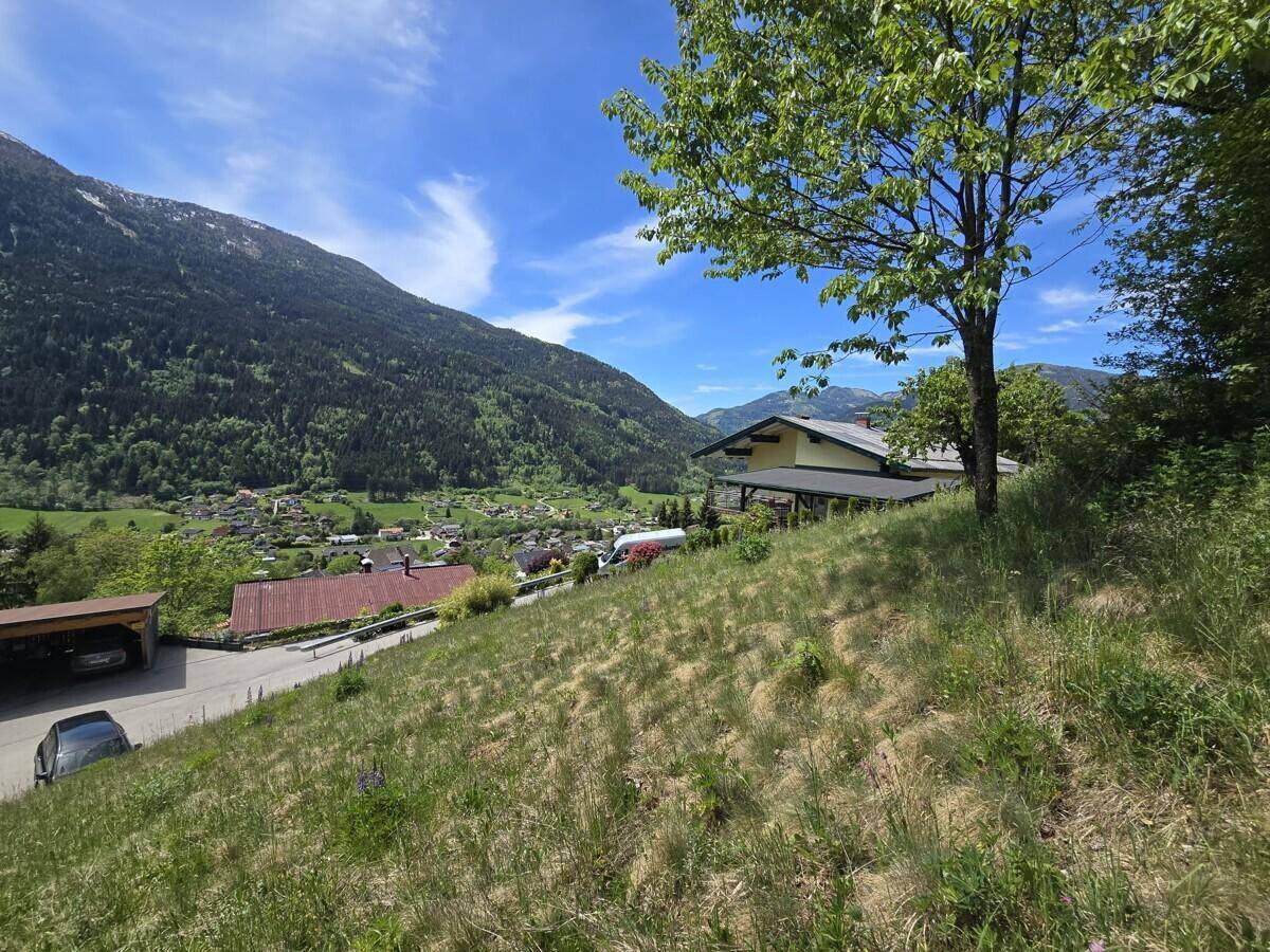 Grüne Hanglage mit einem Haus und einem Carport im Vordergrund, umgeben von Bergen und Natur.