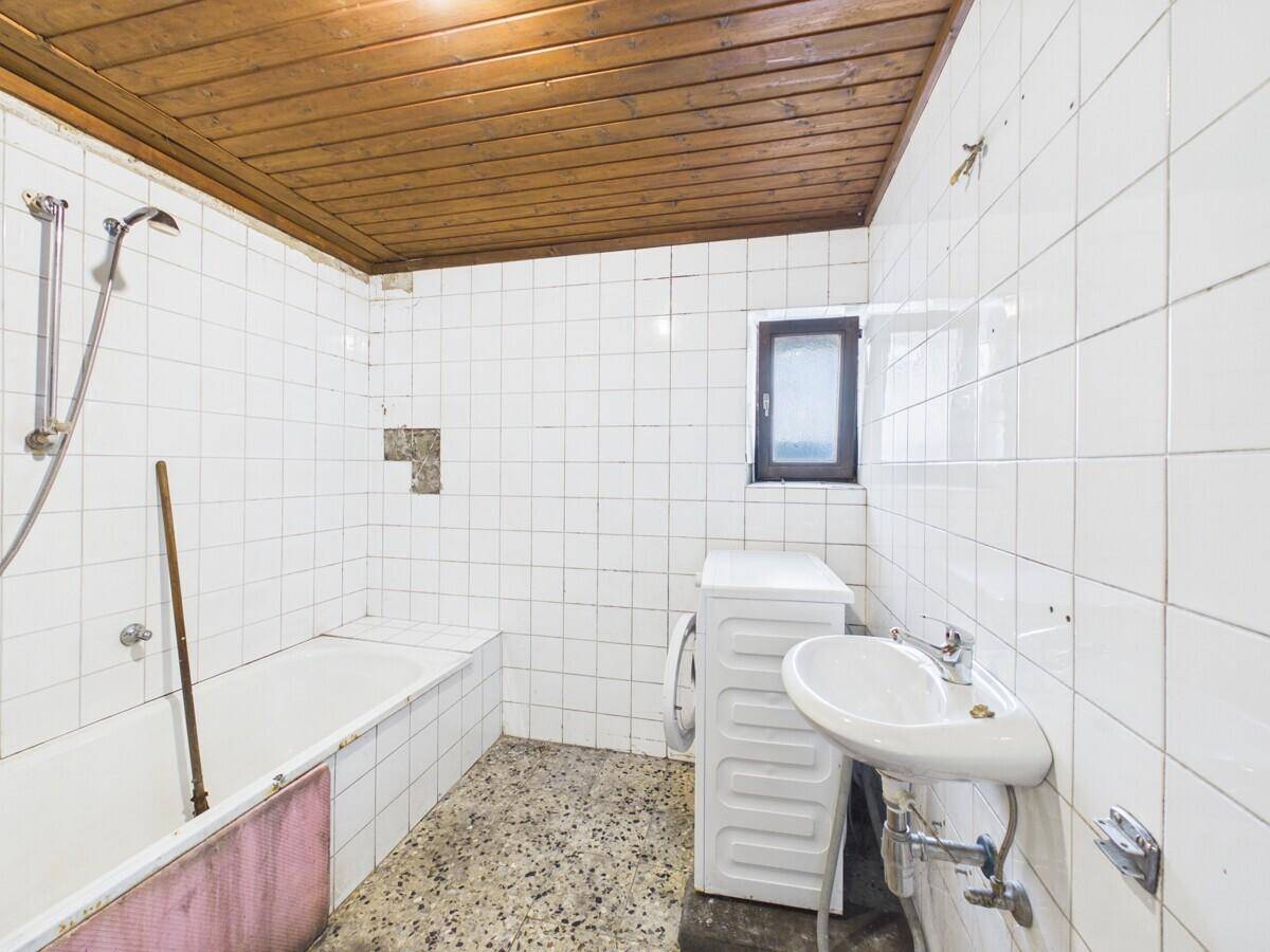 Badezimmer mit weißen Fliesen, Badewanne, Waschbecken und Holzdecke, renovierungsbedürftig.