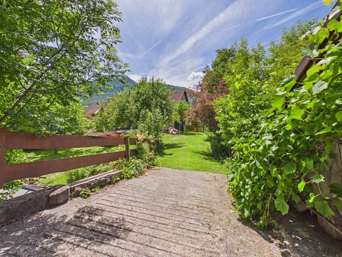 Betonweg führt in einen weitläufigen Gartenbereich mit Bäumen und Sträuchern.