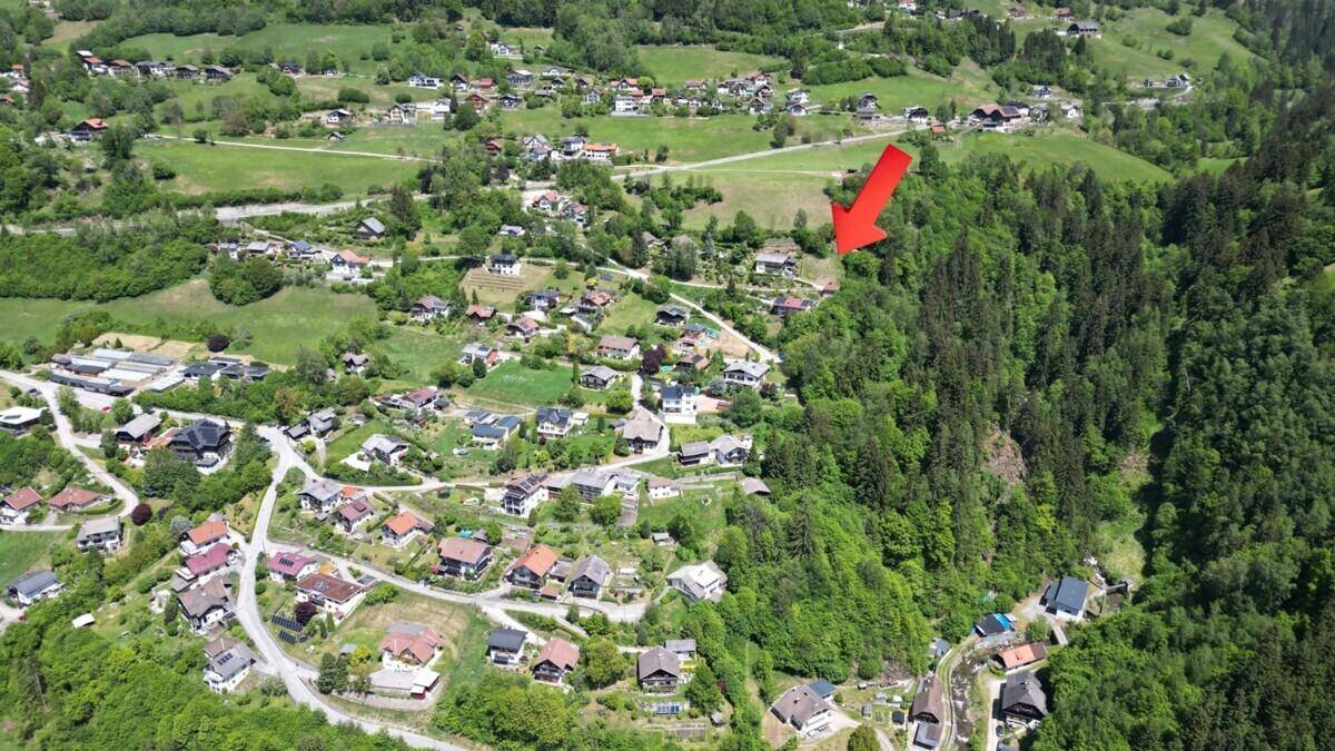 Luftaufnahme eines Dorfes in einem grünen Tal, mit einem roten Pfeil, der auf ein bestimmtes Gebiet zeigt.