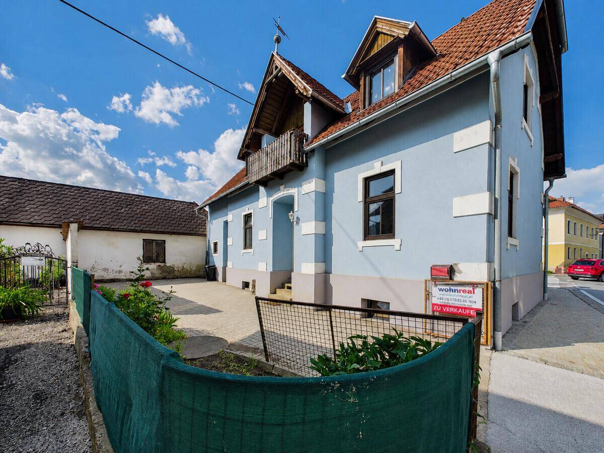 Außenansicht des Hauses von der Straße mit einem "Zu verkaufen"-Schild und grünem Zaun.