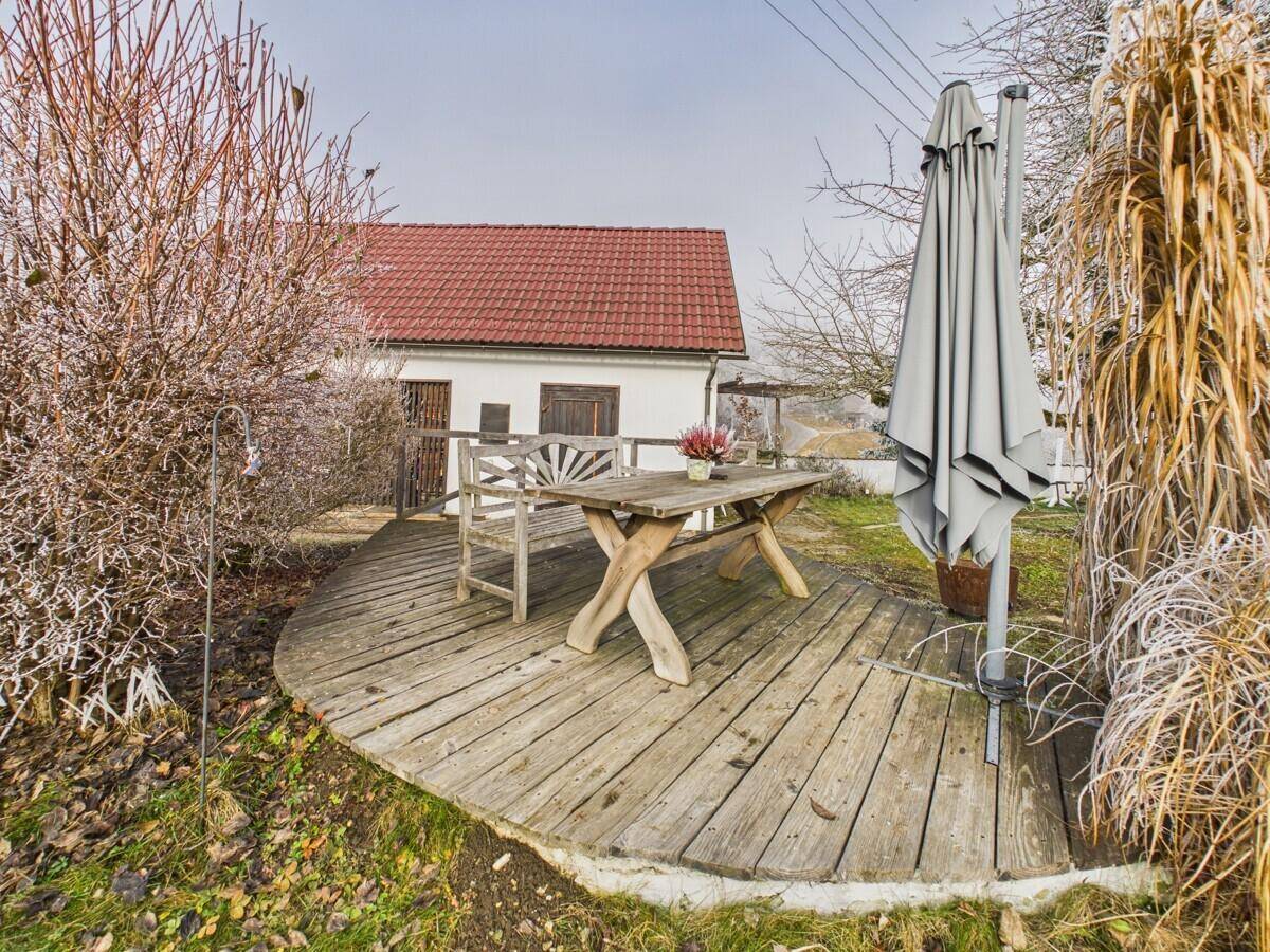 Rustikale Holzterrasse mit Esstisch und Sonnenschirm vor einem kleinen Landhaus.