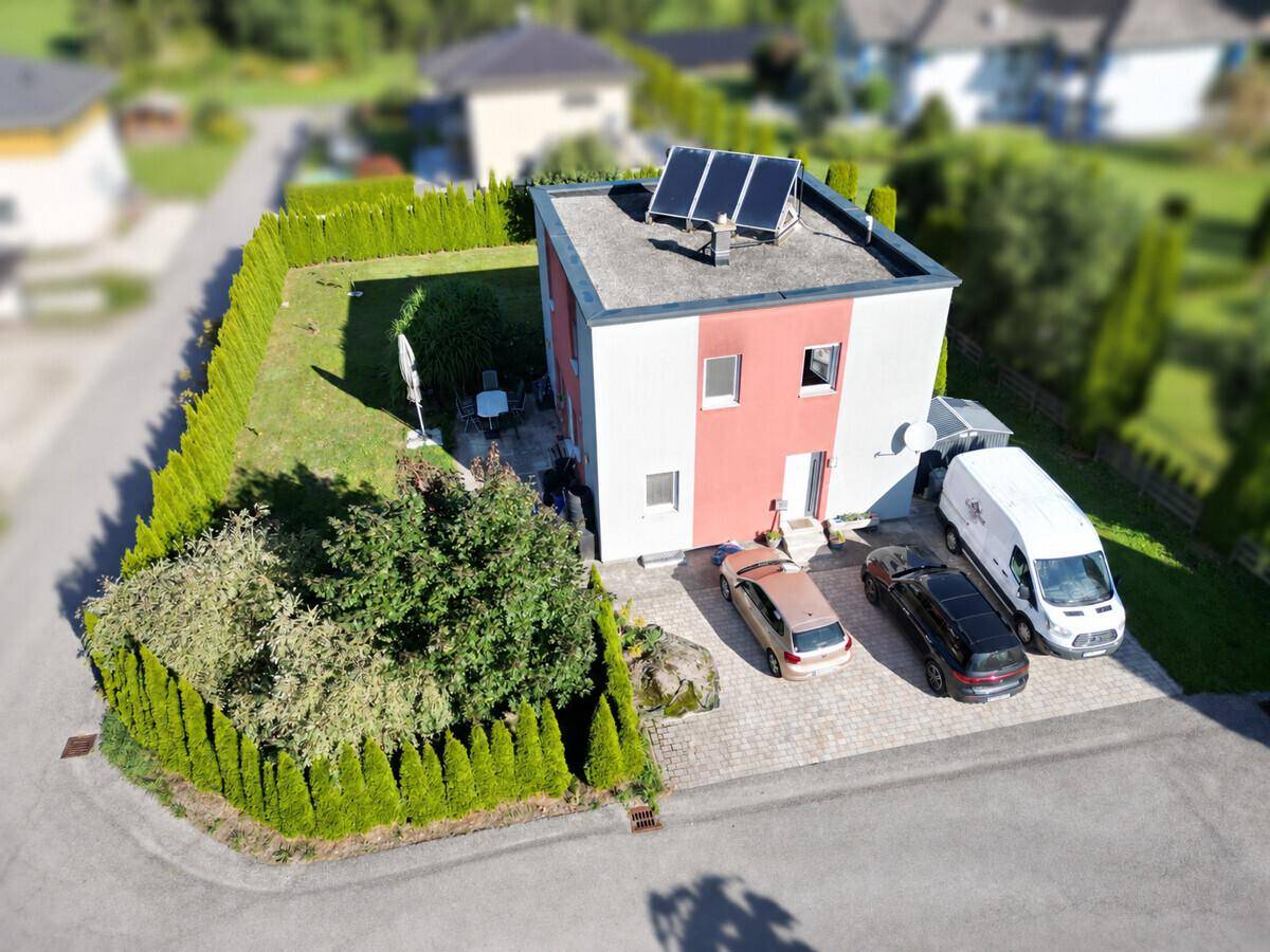 Schrägansicht des Hauses mit Garten, gepflasterter Einfahrt und Photovoltaikanlage auf dem Flachdach.
