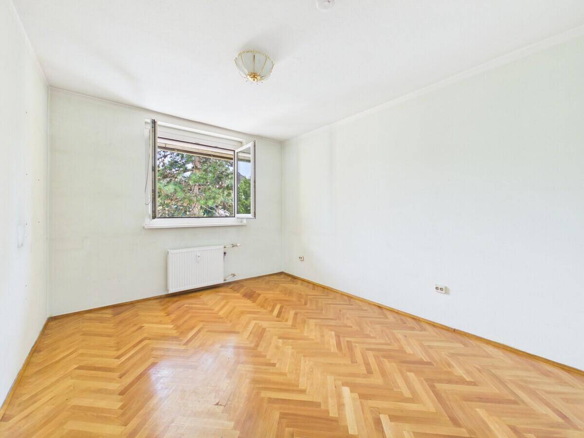 Helles, leeres Zimmer mit Fischgrätparkett und Fenster mit Blick ins Grüne.