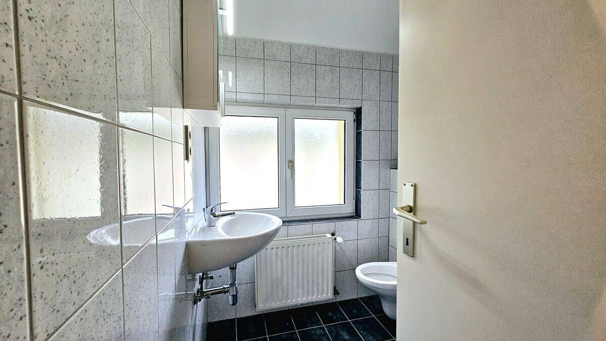 Funktionales Badezimmer mit Waschbecken, Toilette und Fenster, ausgestattet mit hellen Fliesen und Heizkörper.