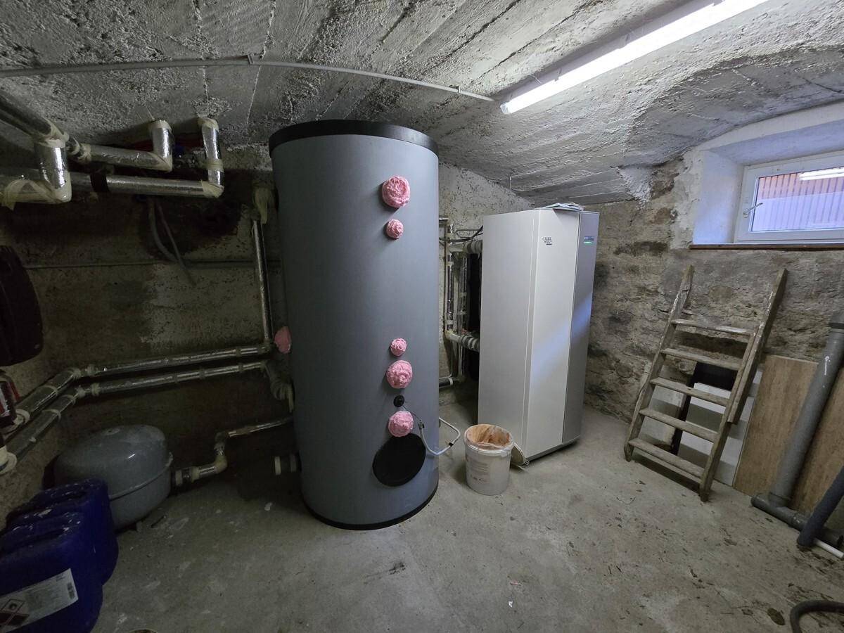 Technikraum im Keller mit Warmwasserspeicher und Wärmepumpe, umgeben von rauen Betonwänden.