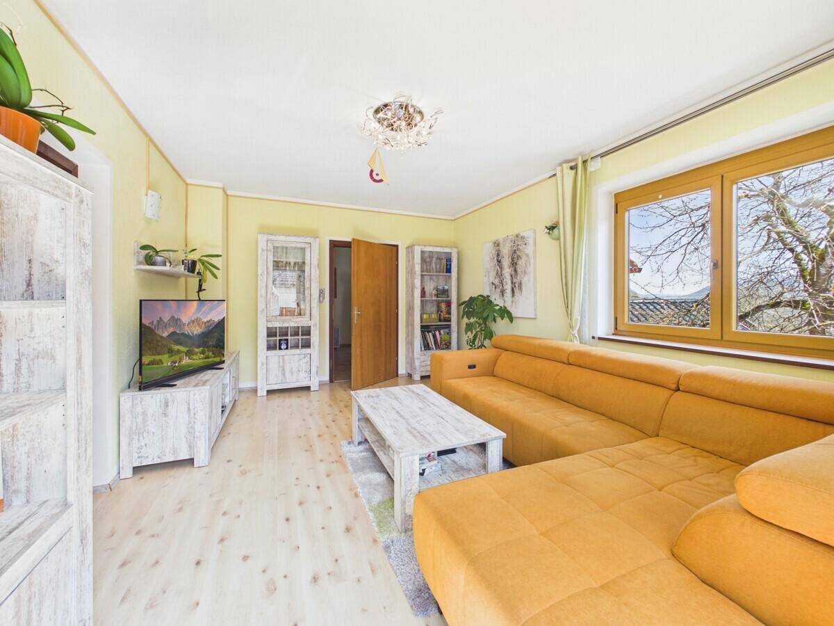 Helles Wohnzimmer mit gelbem Ecksofa, Holzboden und Blick ins Grüne durch das Fenster.