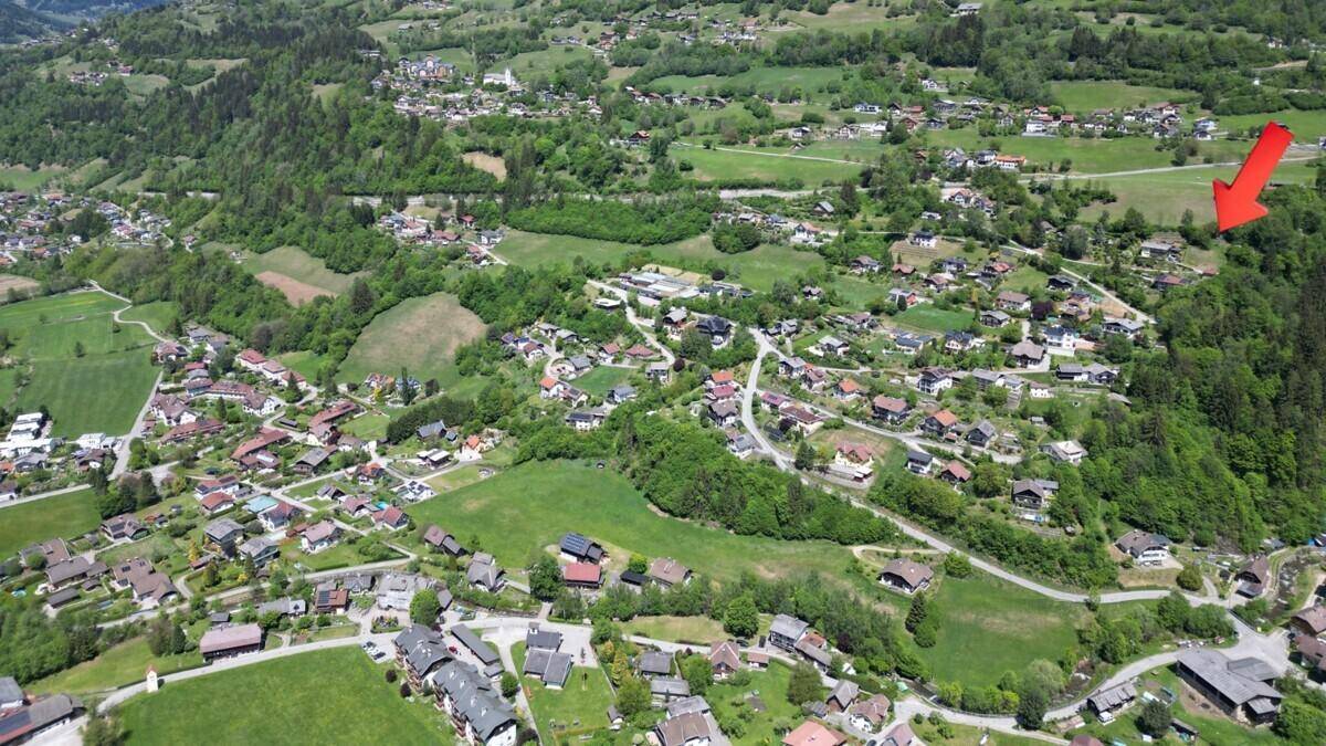 Luftaufnahme eines Dorfes in einem grünen Tal, mit einem roten Pfeil, der auf ein bestimmtes Gebiet zeigt.