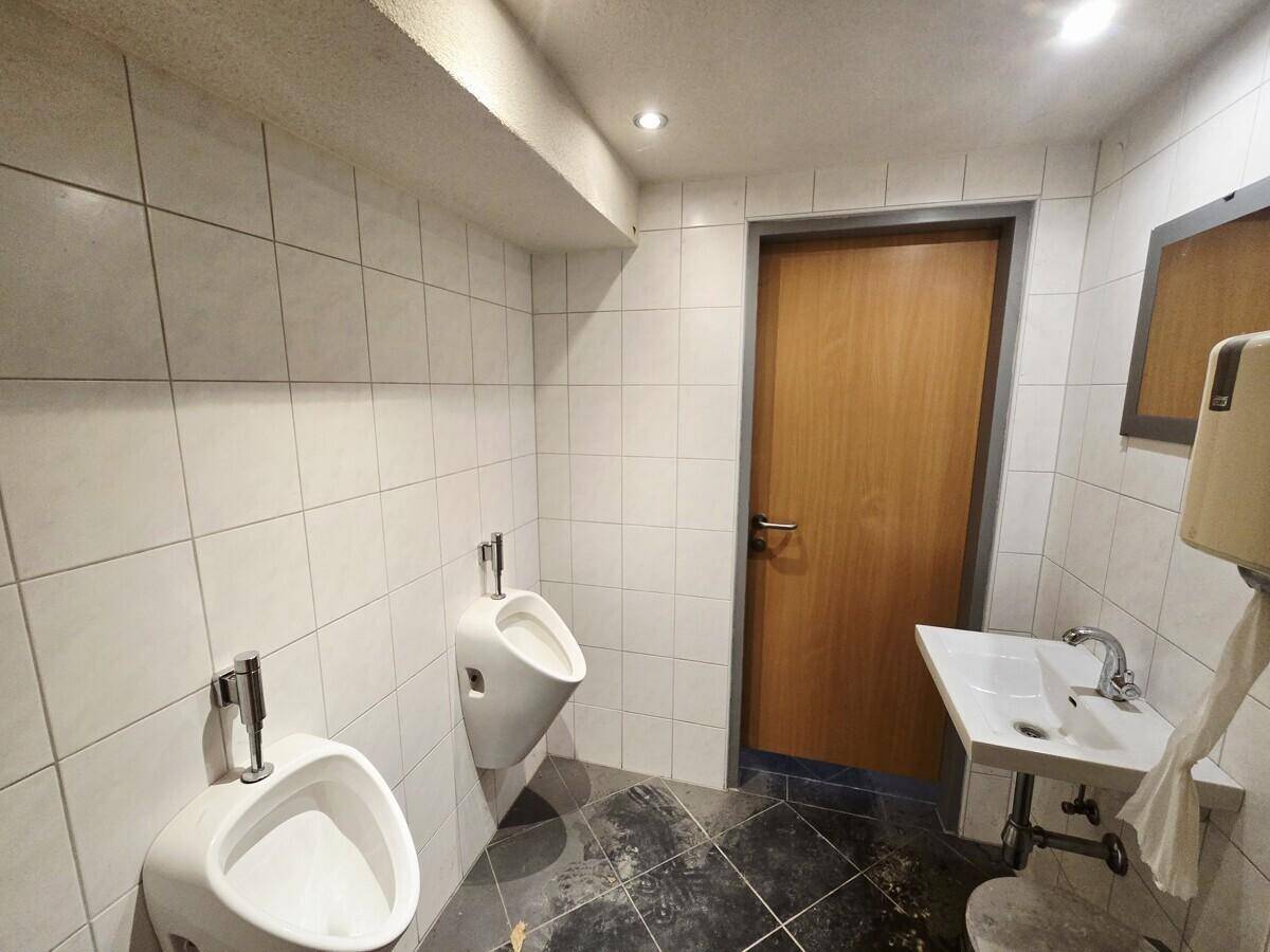 Herren-WC mit zwei Urinalen, einem Waschbecken und weißen Fliesen an den Wänden.