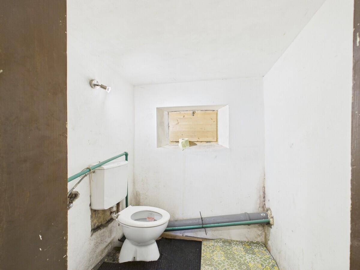 Einfaches WC mit Toilette und sichtbaren Rohrleitungen, das renovierungsbedürftig ist.