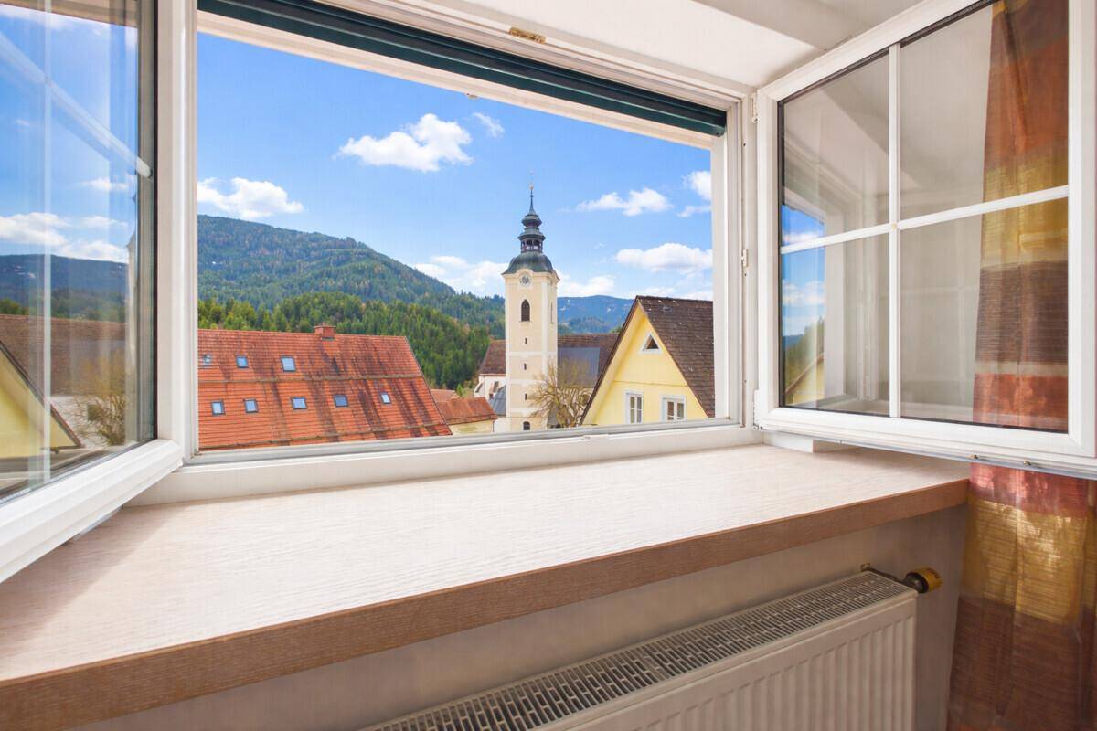 Offenes Fenster mit Blick auf eine Kirche und die umliegende Berglandschaft unter blauem Himmel.