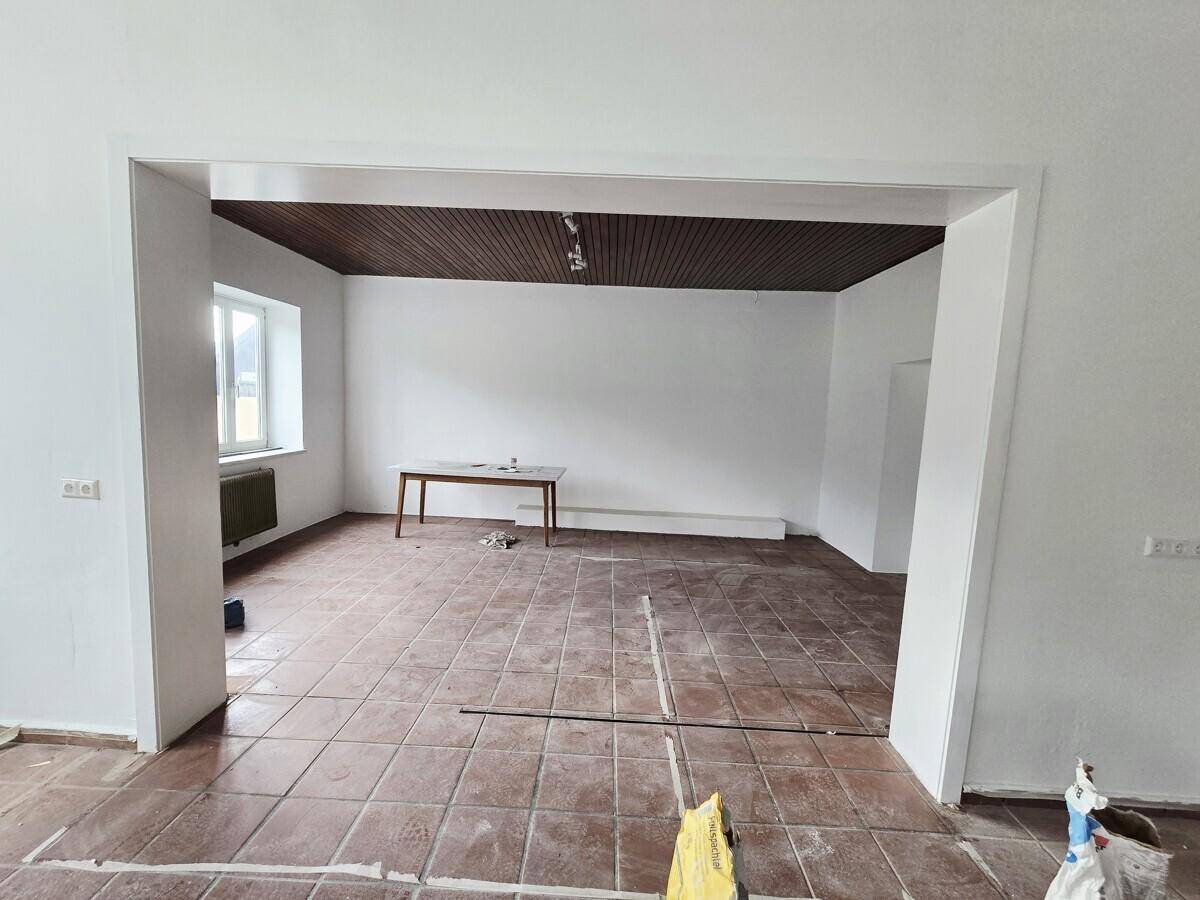 Innenraum mit dunkler Holzdecke und Fliesenboden, der durch einen breiten Durchgang mit einem weiteren Bereich verbunden ist.