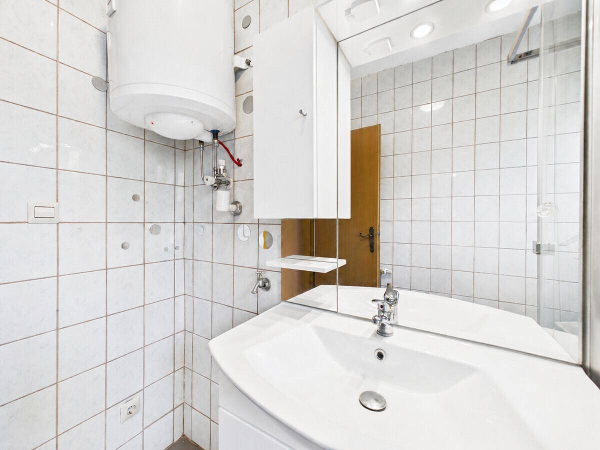 Badezimmer mit weißen Fliesen, Waschbecken, Spiegelschrank und Warmwasserboiler.
