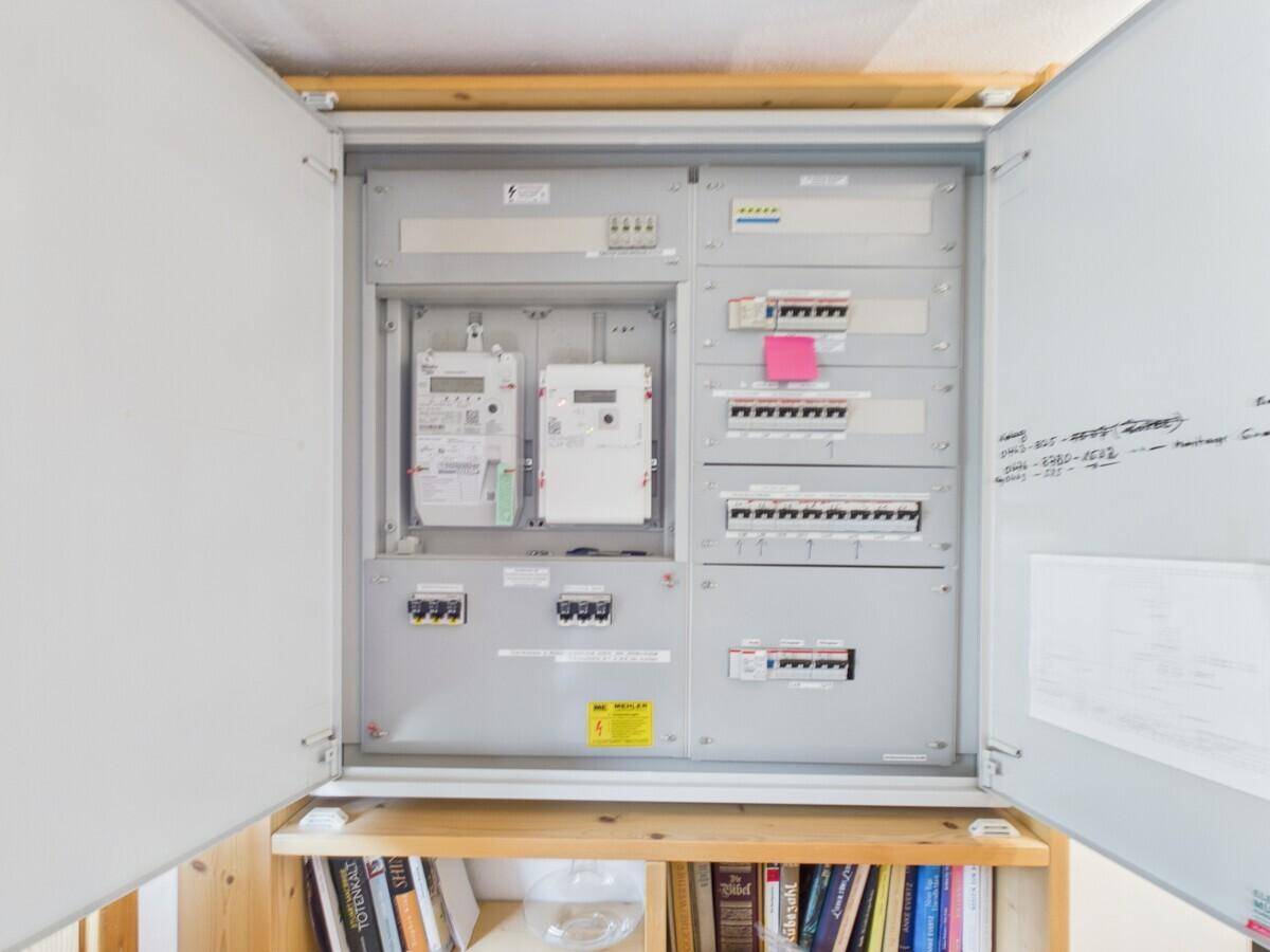Offener Elektroschrank mit Stromzählern und Sicherungen über einem Bücherregal.