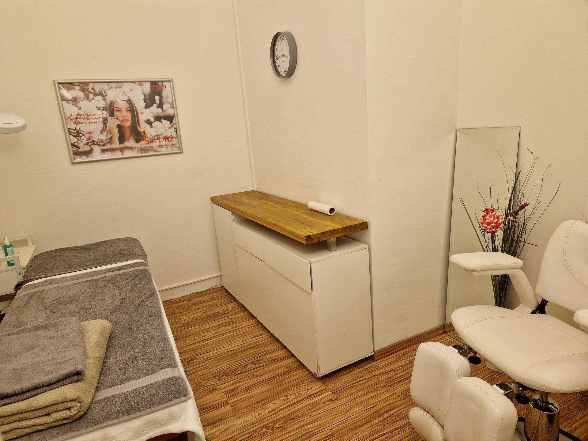 Gemütlicher Behandlungsraum mit Liege, Spiegel und einem kleinen Schrank mit Holzplatte für Utensilien.