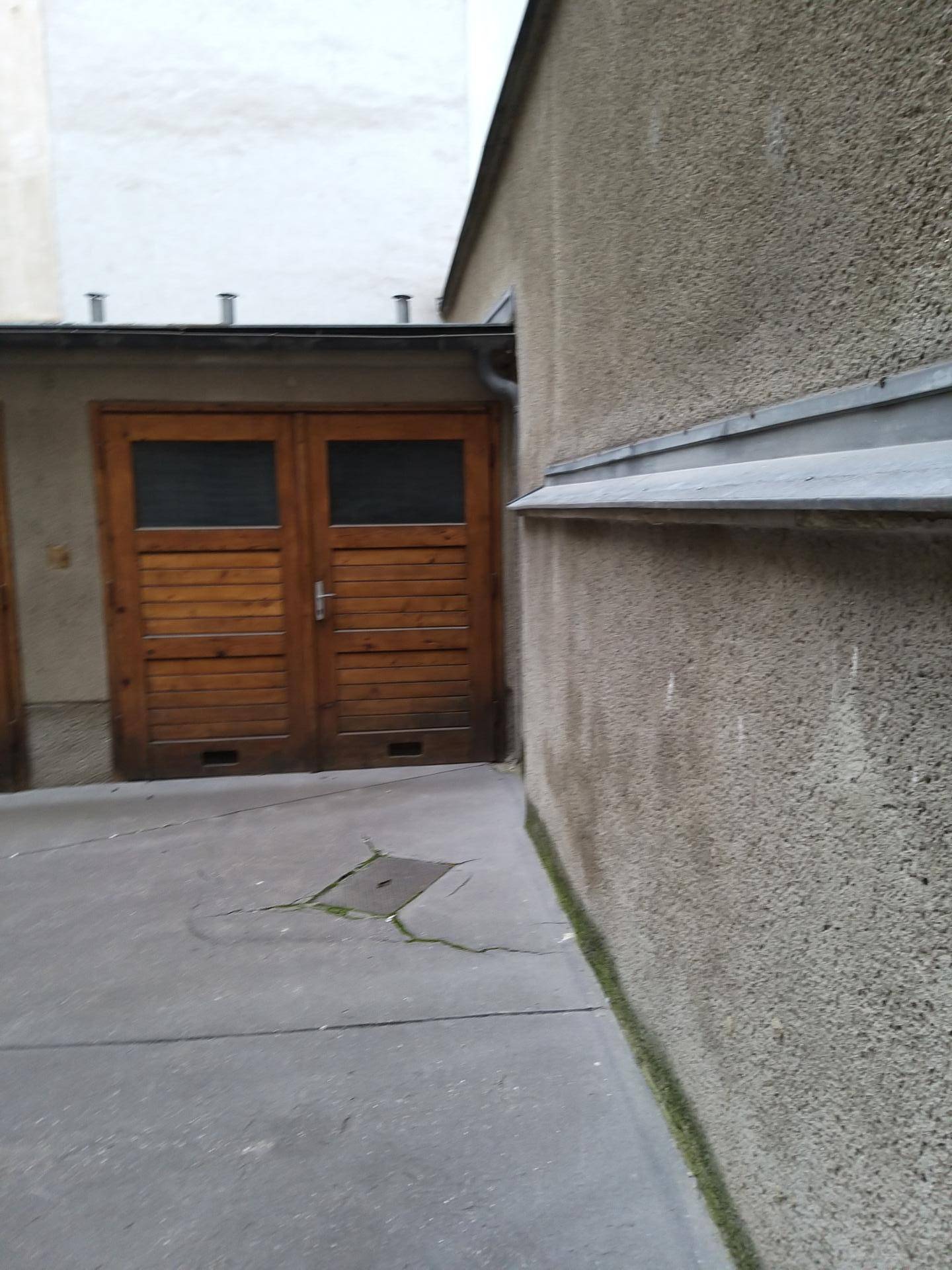 Außenansicht einer Gebäudewand und einer Holztür in einem schmalen Hofbereich mit Betonboden.