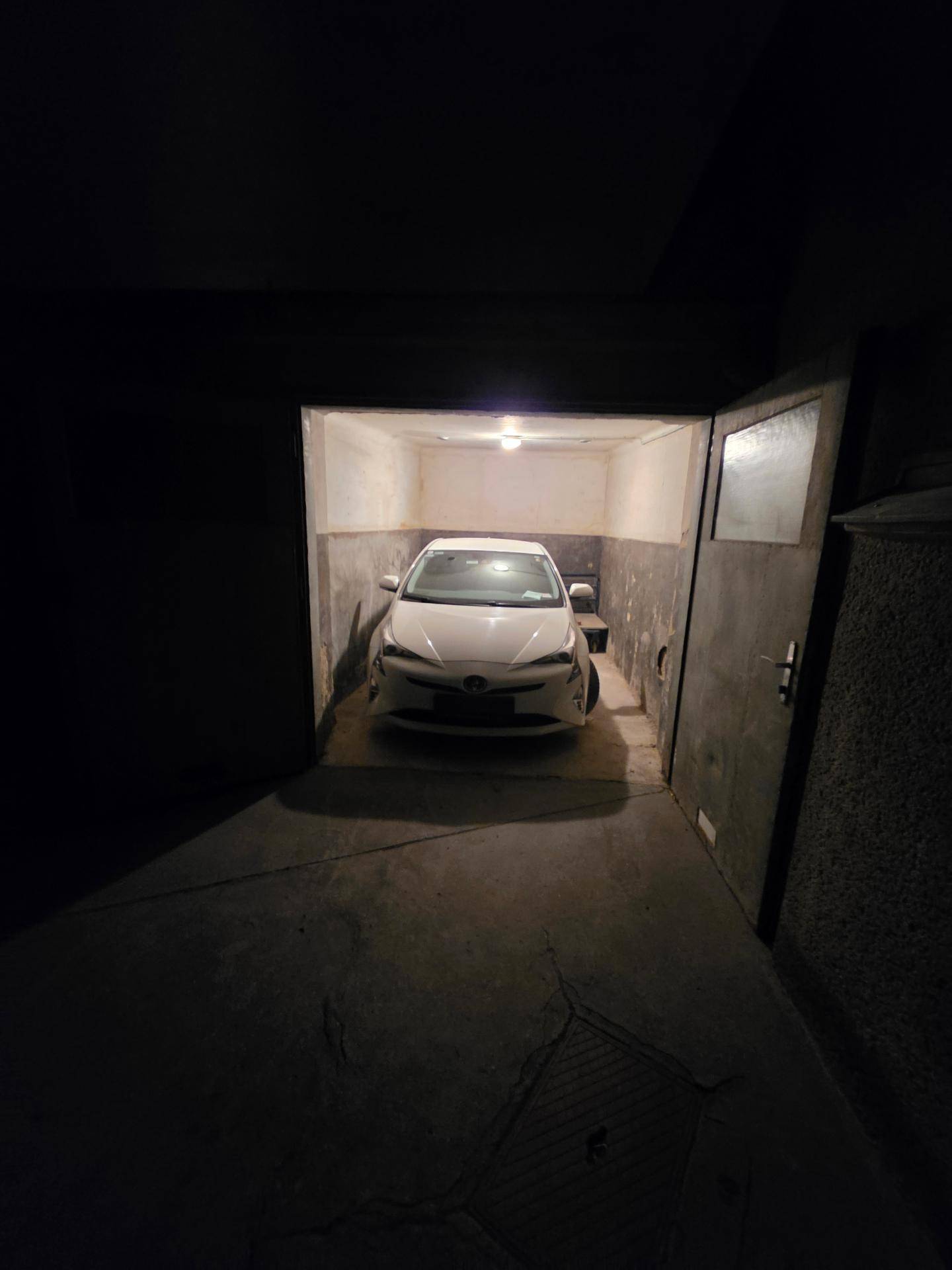 Innenansicht einer Garage mit einem weißen Auto, beleuchtet von einer einzelnen Deckenlampe.