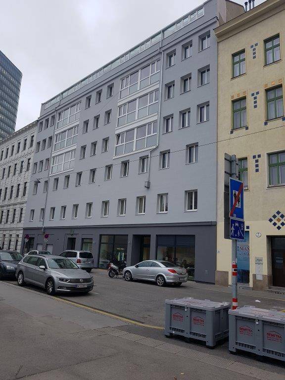 Modernes Mehrfamilienhaus mit grauer Fassade, vielen Fenstern und Geschäften im Erdgeschoss an einer Stadtstraße.