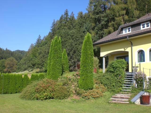 Gelbes Haus mit gepflegtem Garten und hohen Bäumen im Hintergrund unter blauem Himmel.